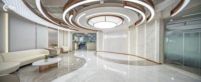 Sảnh chờ Shinhan Life sang trọng với sàn đá trắng bóng loáng, sofa trắng kem mềm mại, đèn trần thiết kế độc đáo và logo Shinhan Life nổi bật trên tường đá.