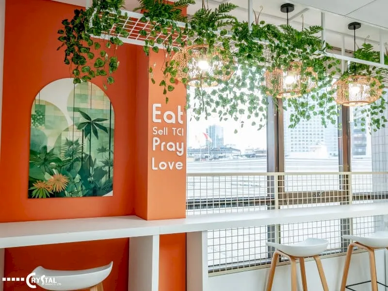 Khu vực pantry văn phòng ấm cúng với tường cam, quầy bar trắng, ghế đẩu chân gỗ, trang trí cây xanh và đèn lồng độc đáo.