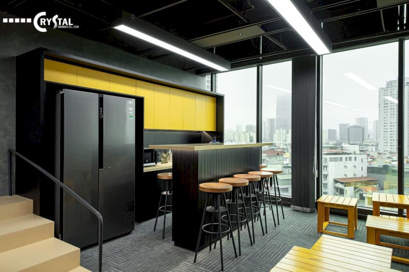 Khu vực bếp văn phòng hiện đại, với tủ lạnh đen, bàn bar và ghế cao, tạo không gian ăn uống và thư giãn thoải mái cho nhân viên.
