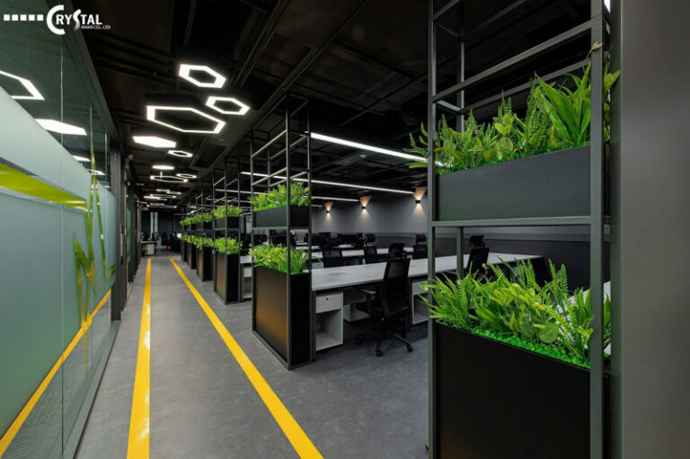 Không gian làm việc tối giản với hàng dài bàn làm việc, được trang trí bằng cây xanh và hệ thống đèn trần hiện đại